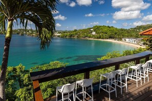 Eine Terrasse mit weißen Korbstühlen mit Blick auf das türkisfarbene Meer und einen Strand.