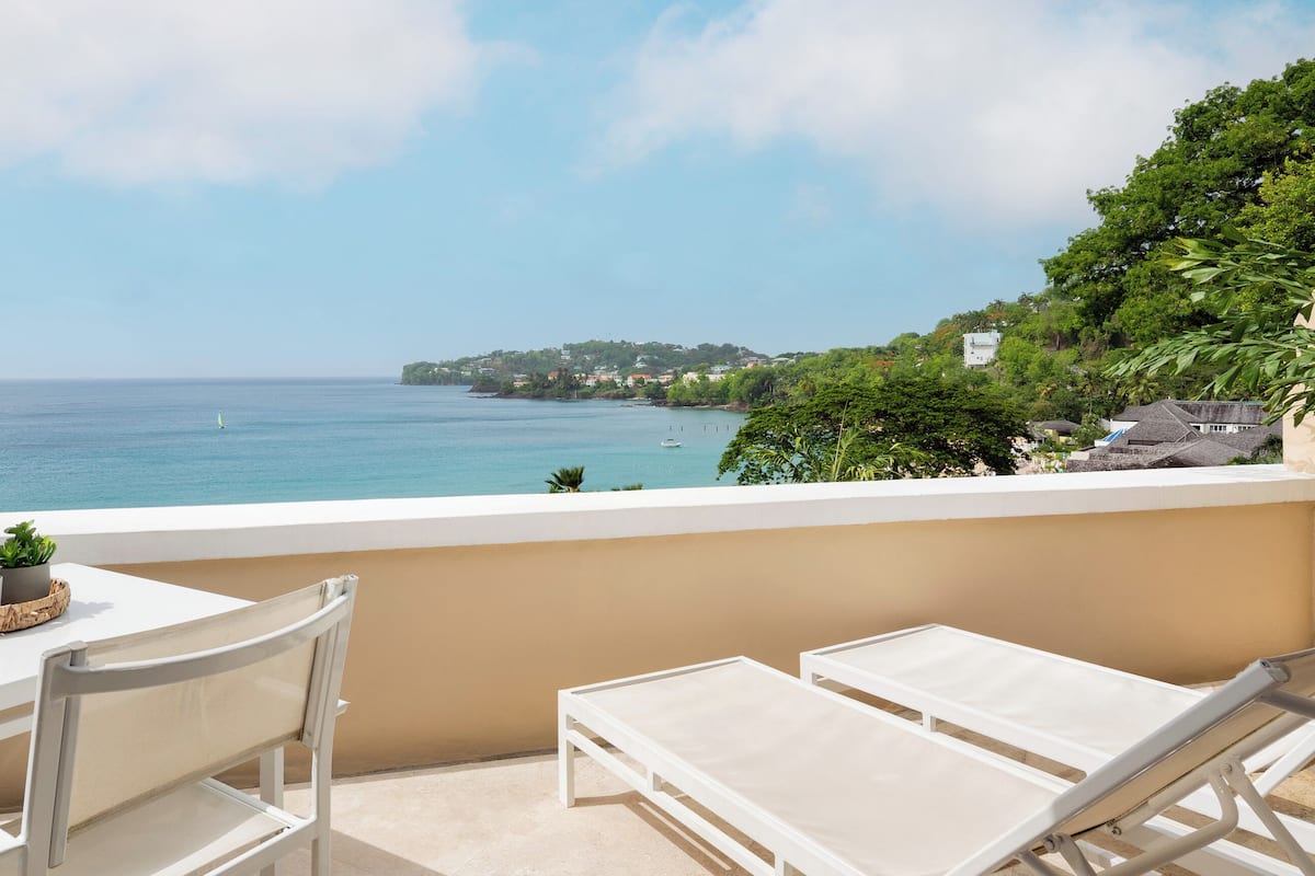 A balcony with a white table and two white lounge chairs, overlooking a coastal view with a clear sky.