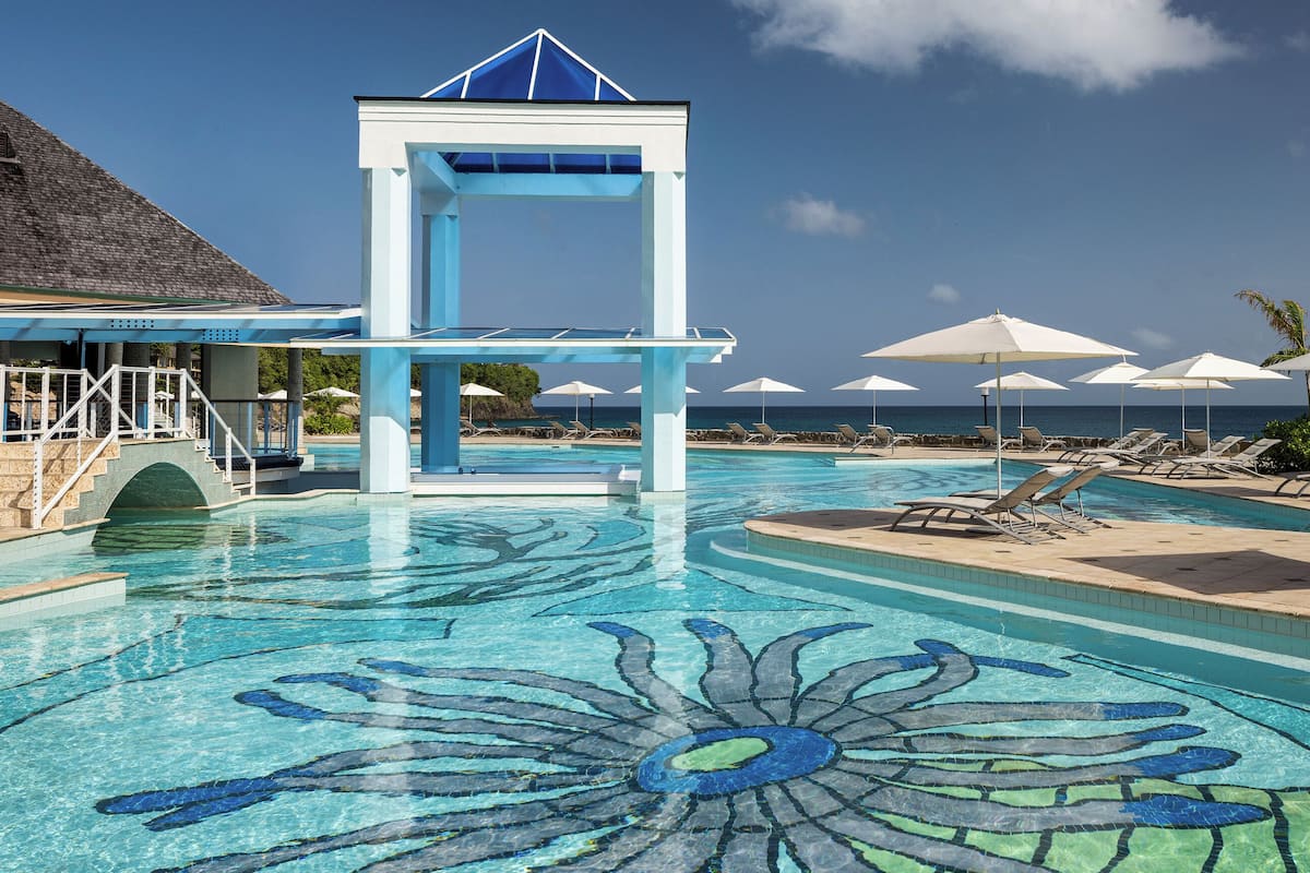 A pool with a blue and white structure, lounge chairs, umbrellas, and a view of the ocean.