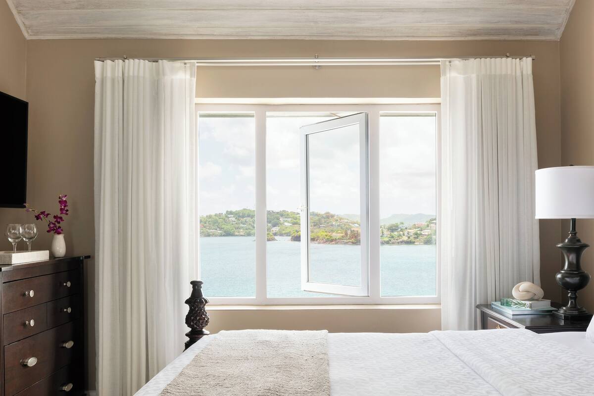 A hotel room with a large bed, a bedside table with a lamp, a dresser, and a window with a view of the sea.