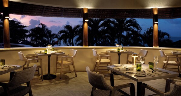 Restaurante com mesas e cadeiras preparadas para refeições, com vista para palmeiras e o oceano ao entardecer.