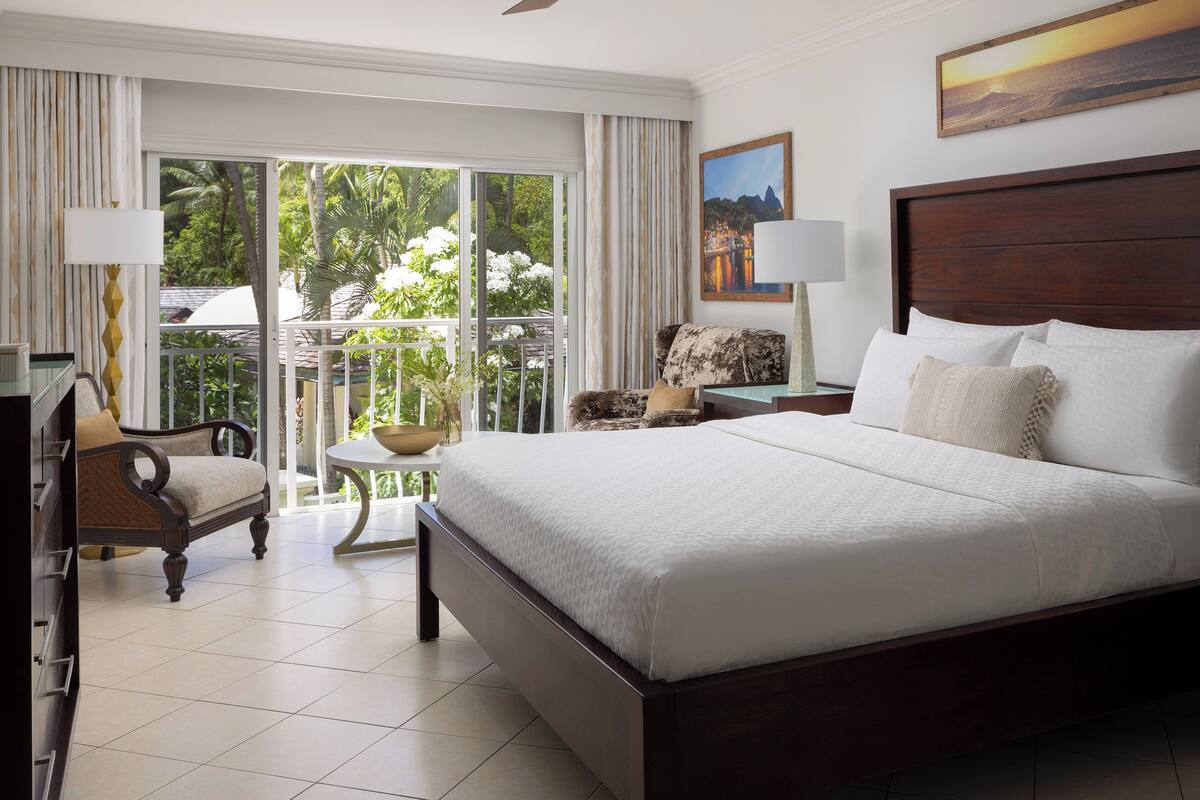 A hotel room with a large bed, two bedside tables, a chair, a small table, and a view of greenery outside.