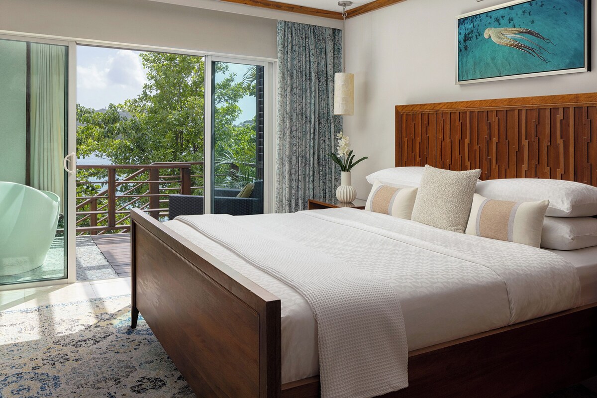 Une chambre à coucher avec une tête de lit en bois, un grand lit avec une literie blanche, une chaise en verre et une vue sur la verdure par une porte-fenêtre coulissante.