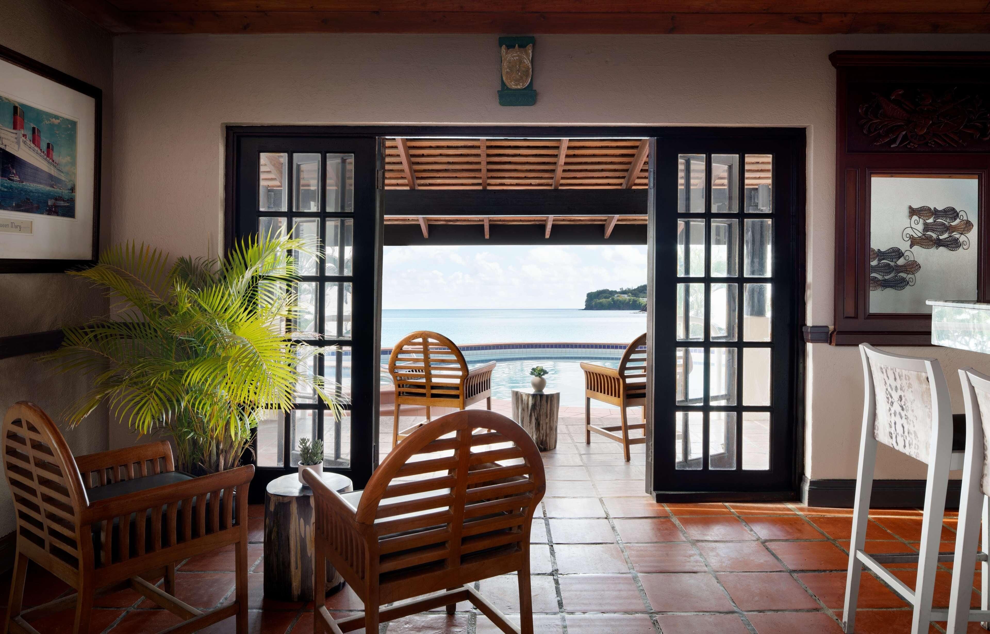 A room with a view of the sea, wooden chairs, a small table, and a potted plant.
