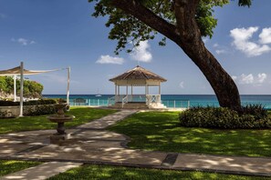 A gazebo with a tiled roof, surrounded by a well-maintained lawn and a pathway leading to the sea. - Sandals Regency La Toc (Castries)