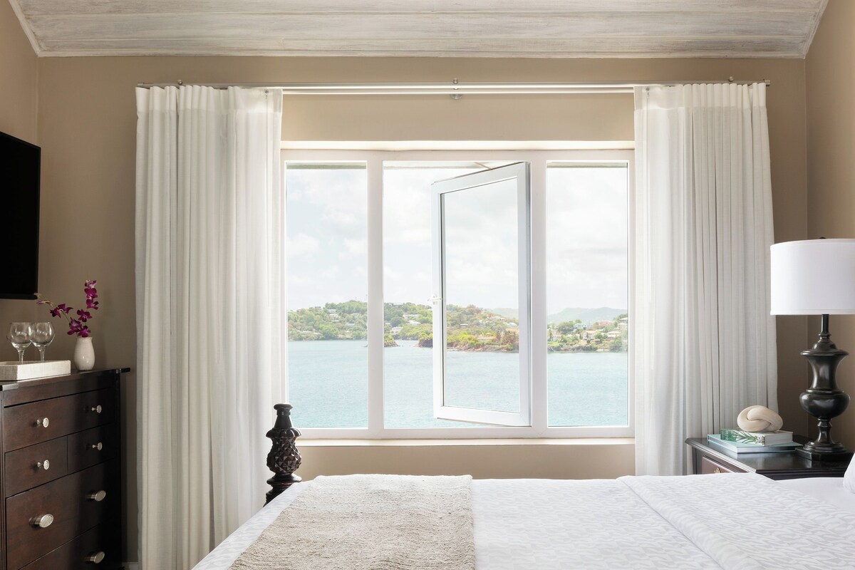Une chambre d’hôtel avec un grand lit, une table de chevet avec une lampe, une commode et une fenêtre donnant sur la mer.