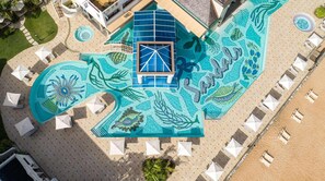 Eine Vogelperspektive zeigt einen luxuriösen Kurort mit einem großen Schwimmbecken, das ein Mosaikdesign aufweist, mehreren Liegebereichen mit Sonnenschirmen und einem Strandabschnitt.