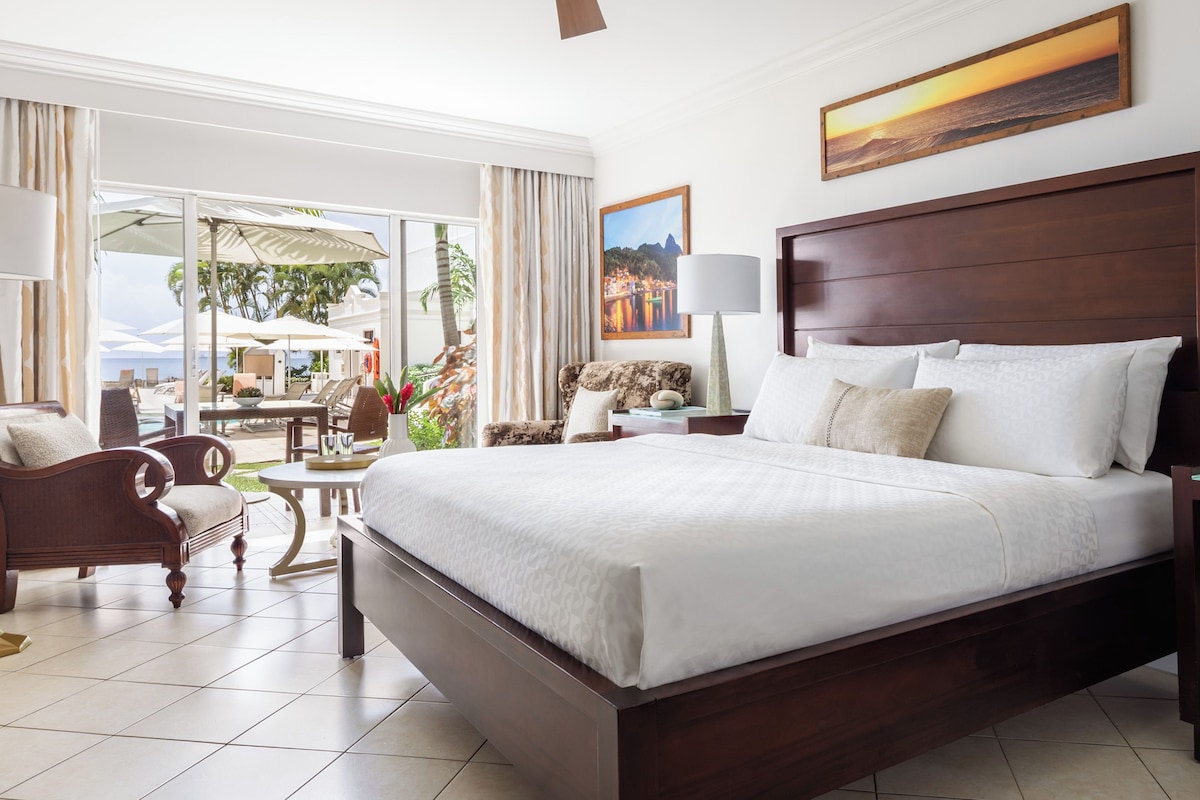 Une chambre spacieuse avec un grand lit, un bureau et une chaise. De grandes fenêtres offrent une vue sur l’extérieur.