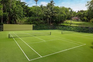 Un court de tennis avec un filet, entouré d’une végétation luxuriante et avec un bâtiment en arrière-plan.