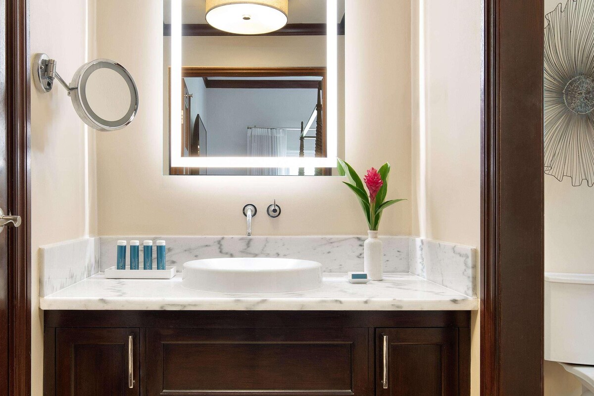 Une salle de bain avec un lavabo en marbre, un miroir, un vase avec une fleur et un élément décoratif mural.