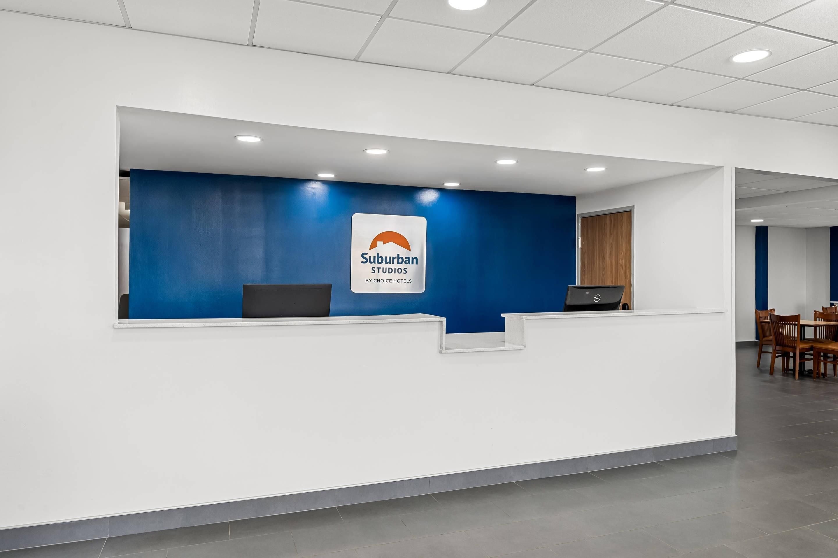 Un mostrador de recepción con una pared azul decorativa, una computadora portátil y un cartel de Suburban Studios by Choice Hotels.