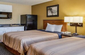 A hotel room with two beds, a kitchenette, a refrigerator, a bedside table with a lamp, and a framed picture on the wall. - Suburban Studios Chester I-95 (Chester)
