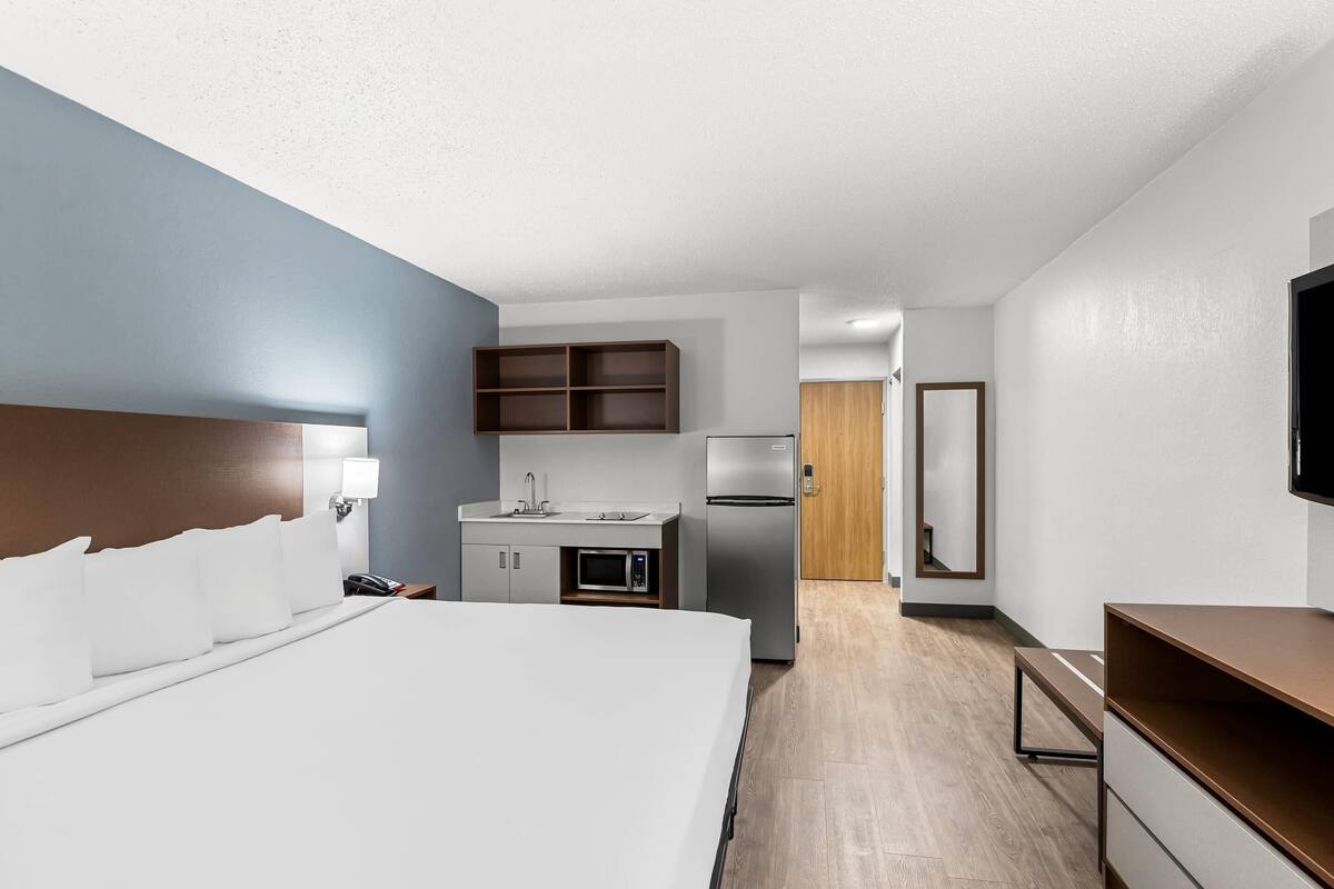 A hotel room with a large bed, a kitchenette with a microwave, refrigerator, and wooden cabinets, a TV mounted on the wall, and a wooden door leading to another room.