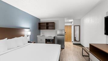 A hotel room with a large bed, a kitchenette with a microwave, refrigerator, and wooden cabinets, a TV mounted on the wall, and a wooden door leading to another room.