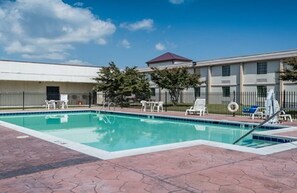 A swimming pool with lounge chairs and a covered area, surrounded by a building with a red roof and a fence. - Suburban Studios Chester I-95 (Chester)