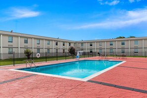 Una piscina con borde de baldosas rojas, rodeada por una cerca, y un edificio con varias ventanas al fondo.