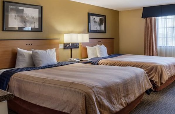 A hotel room with two beds, a headboard, a nightstand, a lamp, and two framed pictures on the wall. - Suburban Studios Chester I-95 (Chester)