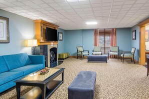 A hotel room with a blue sofa, a small table with a lamp, a television, a fireplace, and a patterned carpet. - Suburban Studios Chester I-95 (Chester)