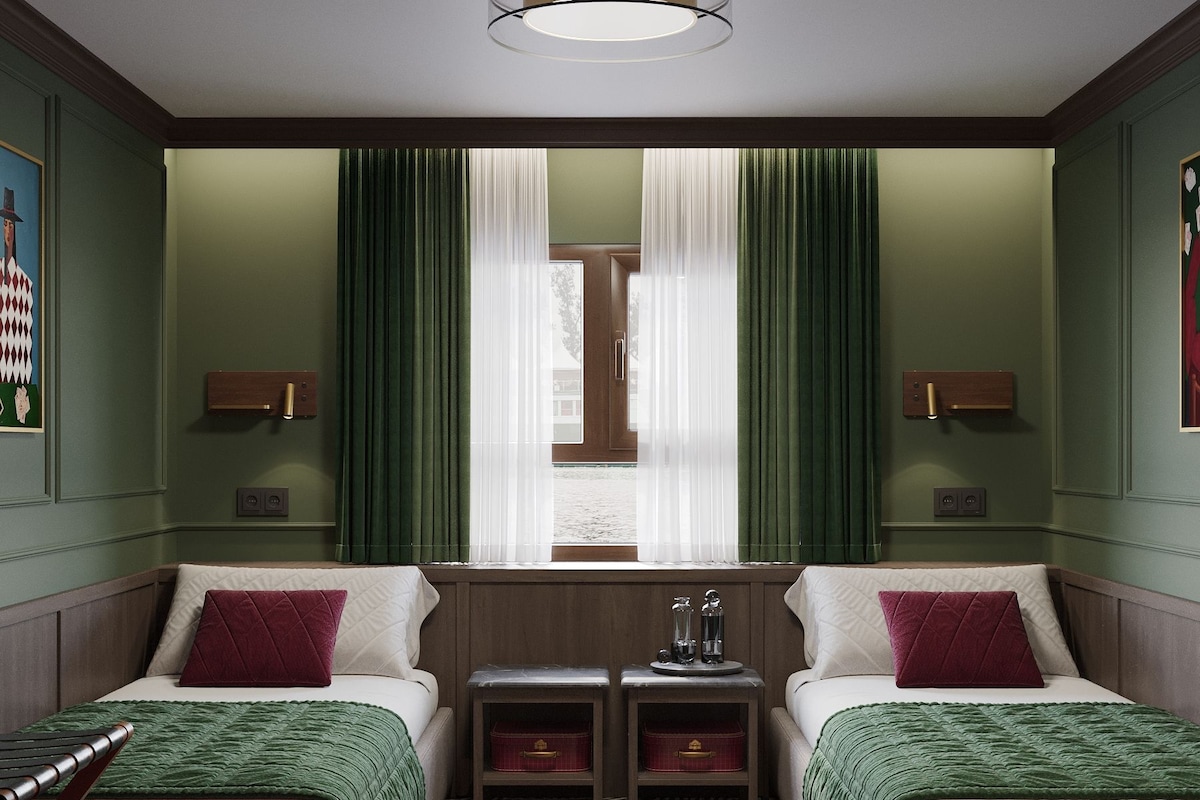 Una camera d'albergo con due letti, una finestra con tende, un piccolo tavolino con una lampada e un pavimento decorato.