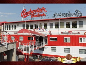 Un bateau rouge et blanc arborant l’inscription « Budweiser Budvar », amarré à un quai où est affiché un panneau indiquant « Hôtel Albatros ».