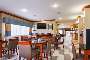 Un'area ristorazione di un hotel con diversi tavoli e sedie, un televisore a schermo piatto montato a parete e vista all'esterno attraverso ampie finestre.
