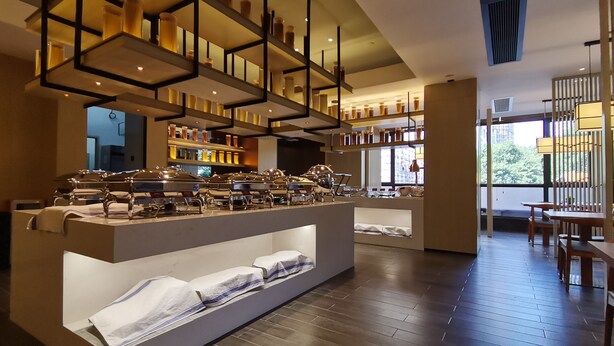 A modern hotel lobby with a central buffet area, wooden flooring, and a view of the outdoors through large windows.