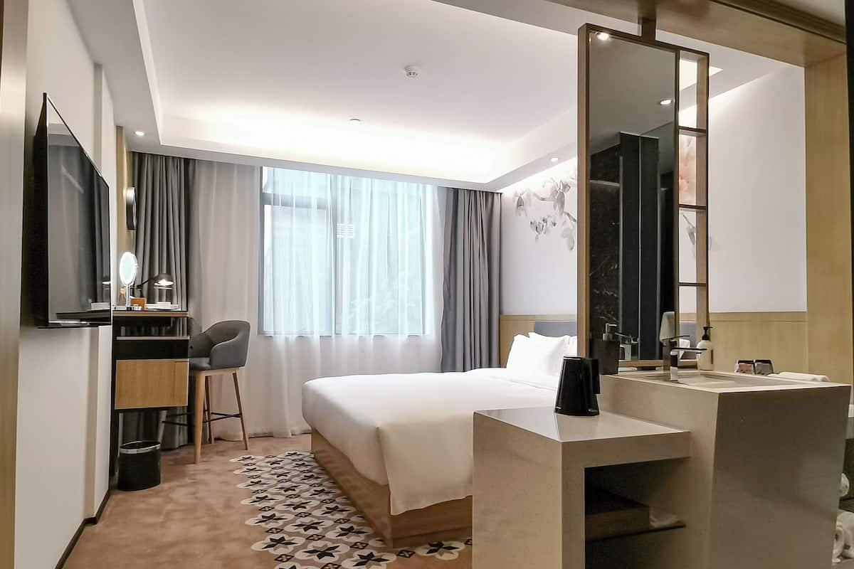 A hotel room with a bed, a desk with a chair, a large mirror, and a bathroom with a sink and mirror.