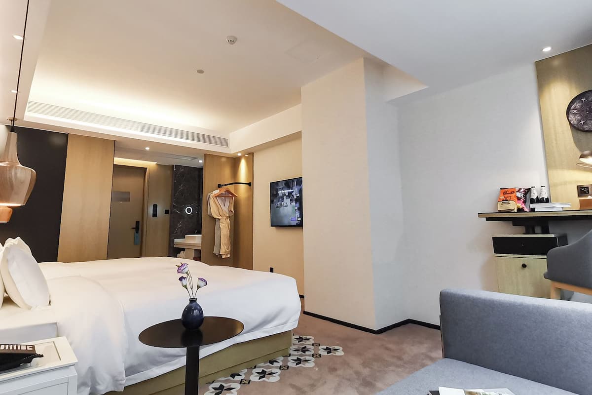 A hotel room with a bed, bedside table, a small round table with a vase, a grey armchair, a kitchenette area, and a bathroom visible through an open door.