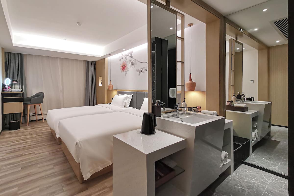 A hotel room with two beds, a desk with a chair, a large mirror, a vanity with a sink, and a decorative wall piece.