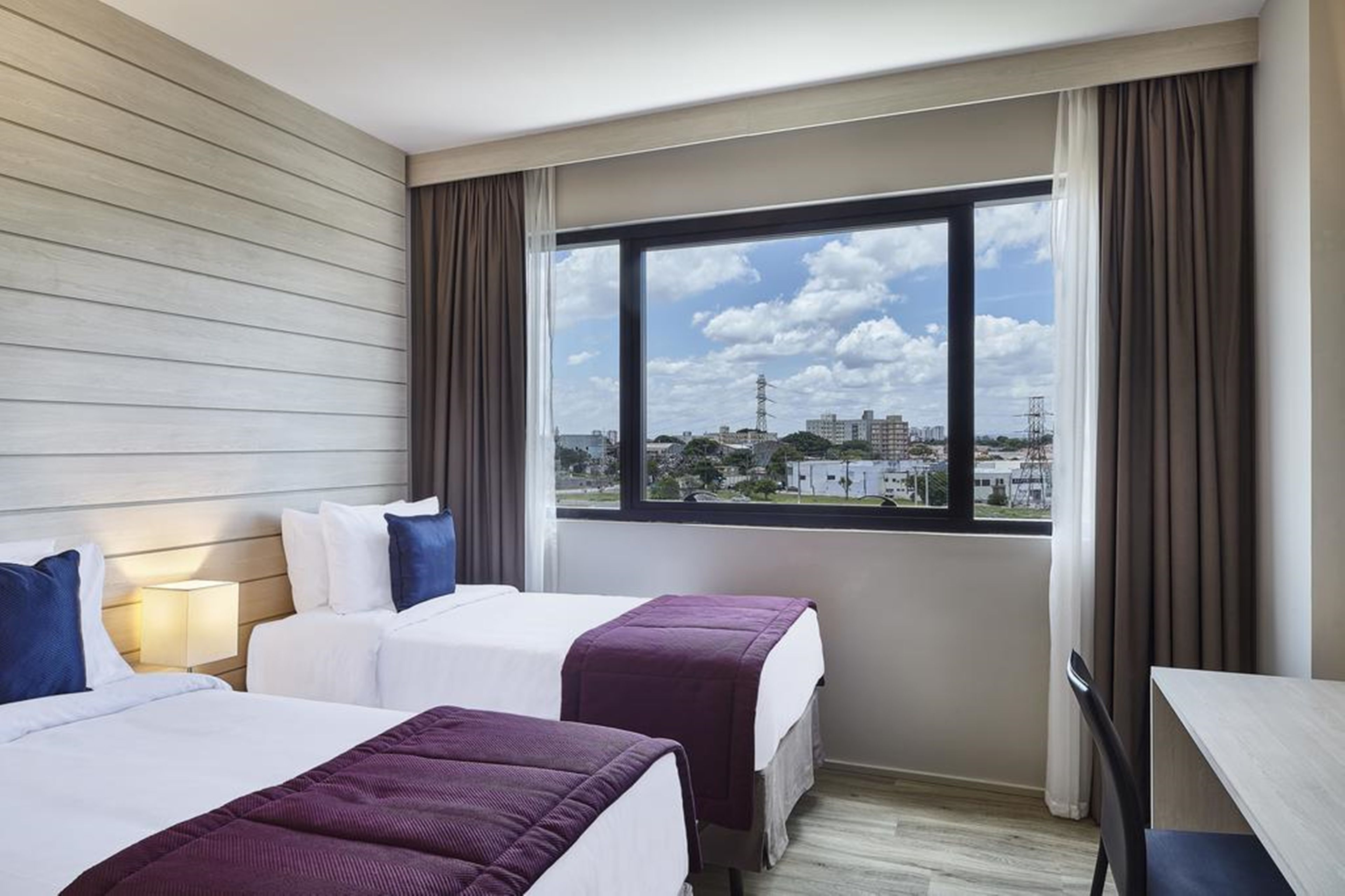 Habitación de hotel con dos camas, un escritorio y un ventanal grande con vistas a la ciudad.
