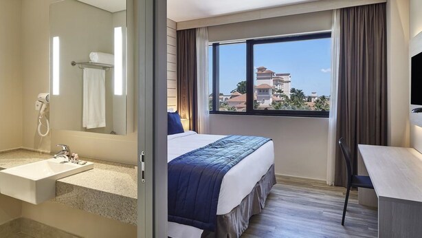 A hotel room with a large bed, a bathroom with a sink and mirror, and a view of buildings through a window. - Contemporaneo (Campinas)
