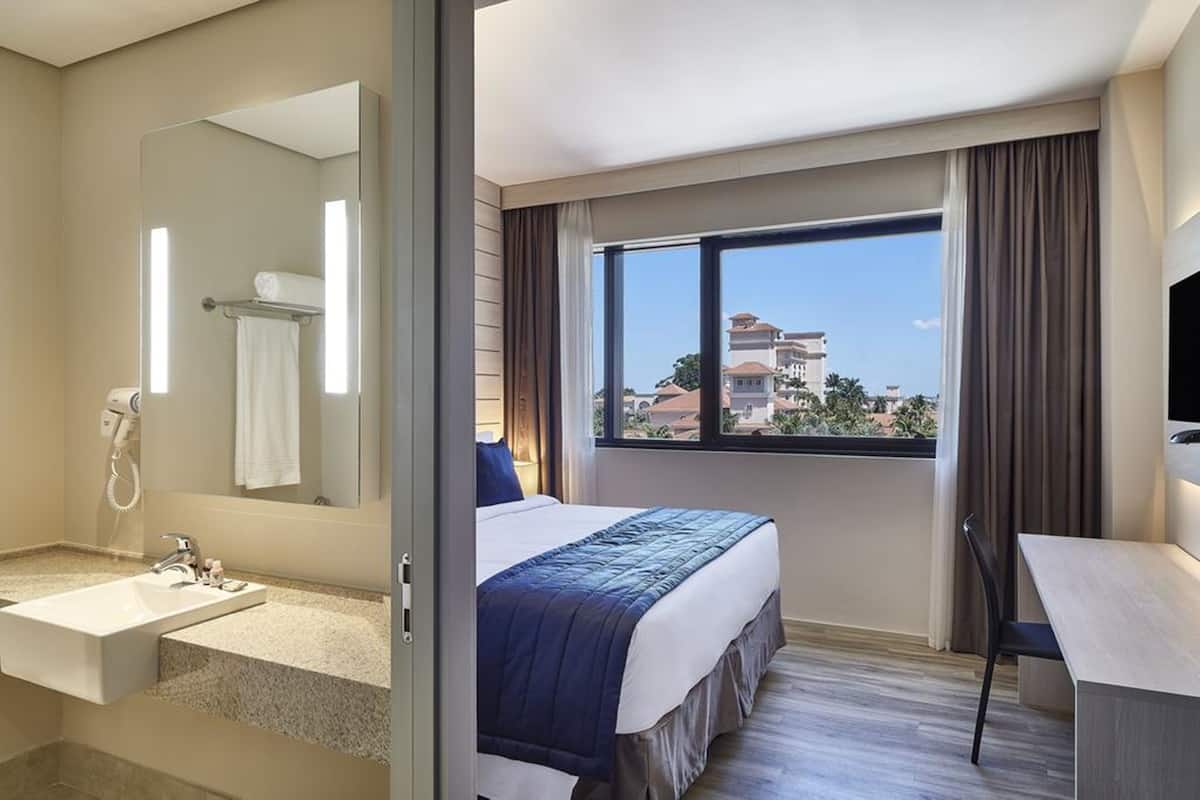 A hotel room with a large bed, a bathroom with a sink and mirror, and a view of buildings through a window.