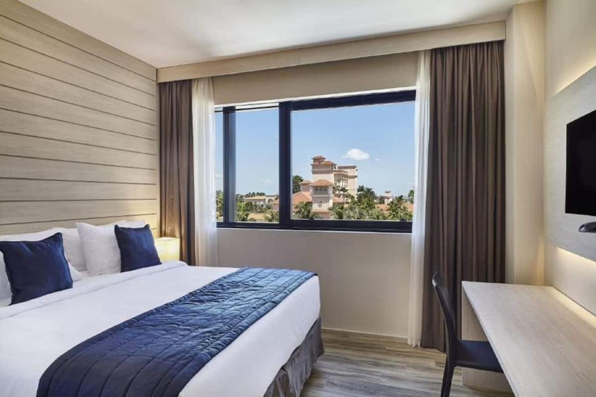 A hotel room with a bed, a desk, and a large window offering a view of buildings and greenery.