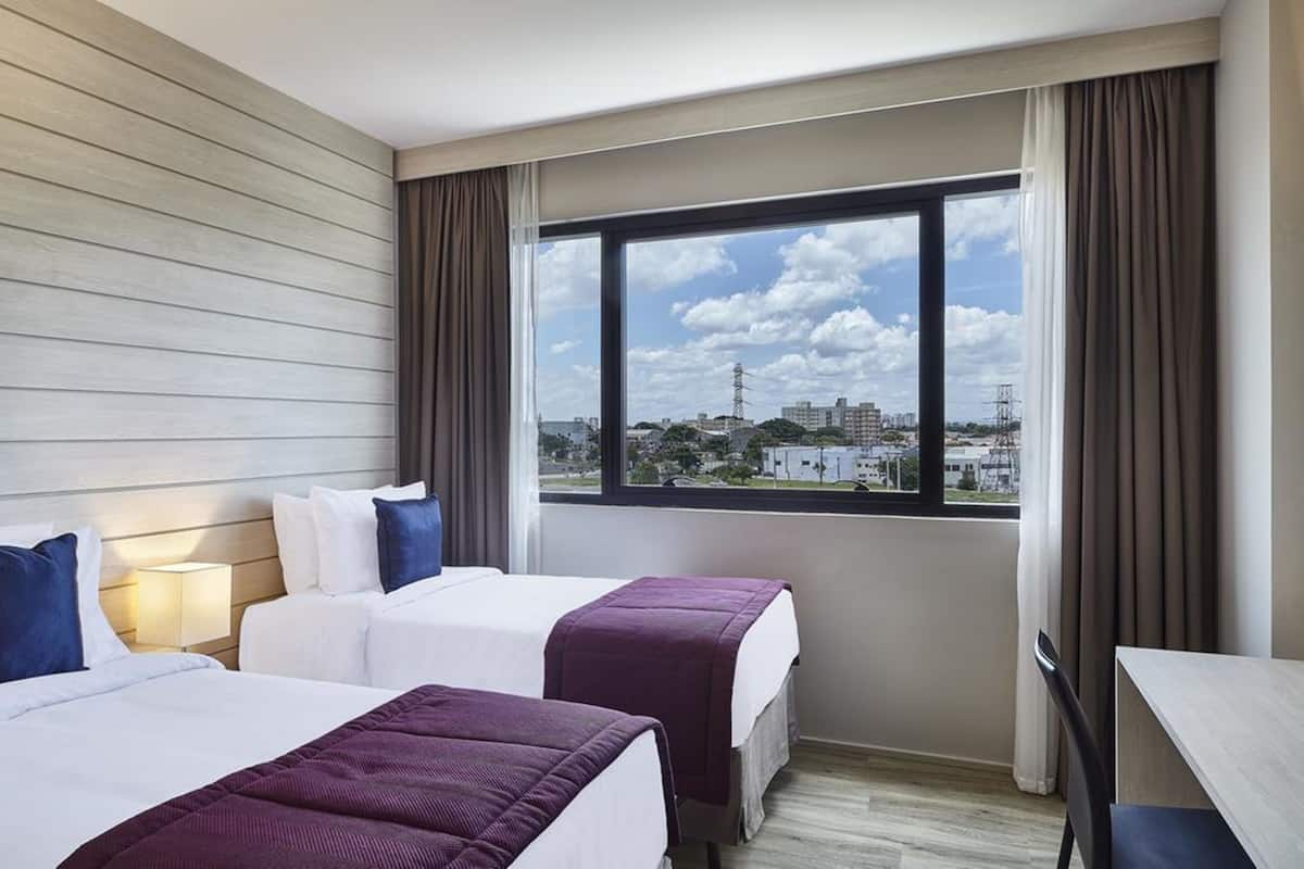 A hotel room with two beds, a desk, and a large window offering a city view.