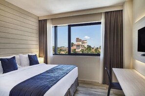 Habitación de hotel con cama, escritorio y ventana grande que ofrece vistas a edificios y áreas verdes.