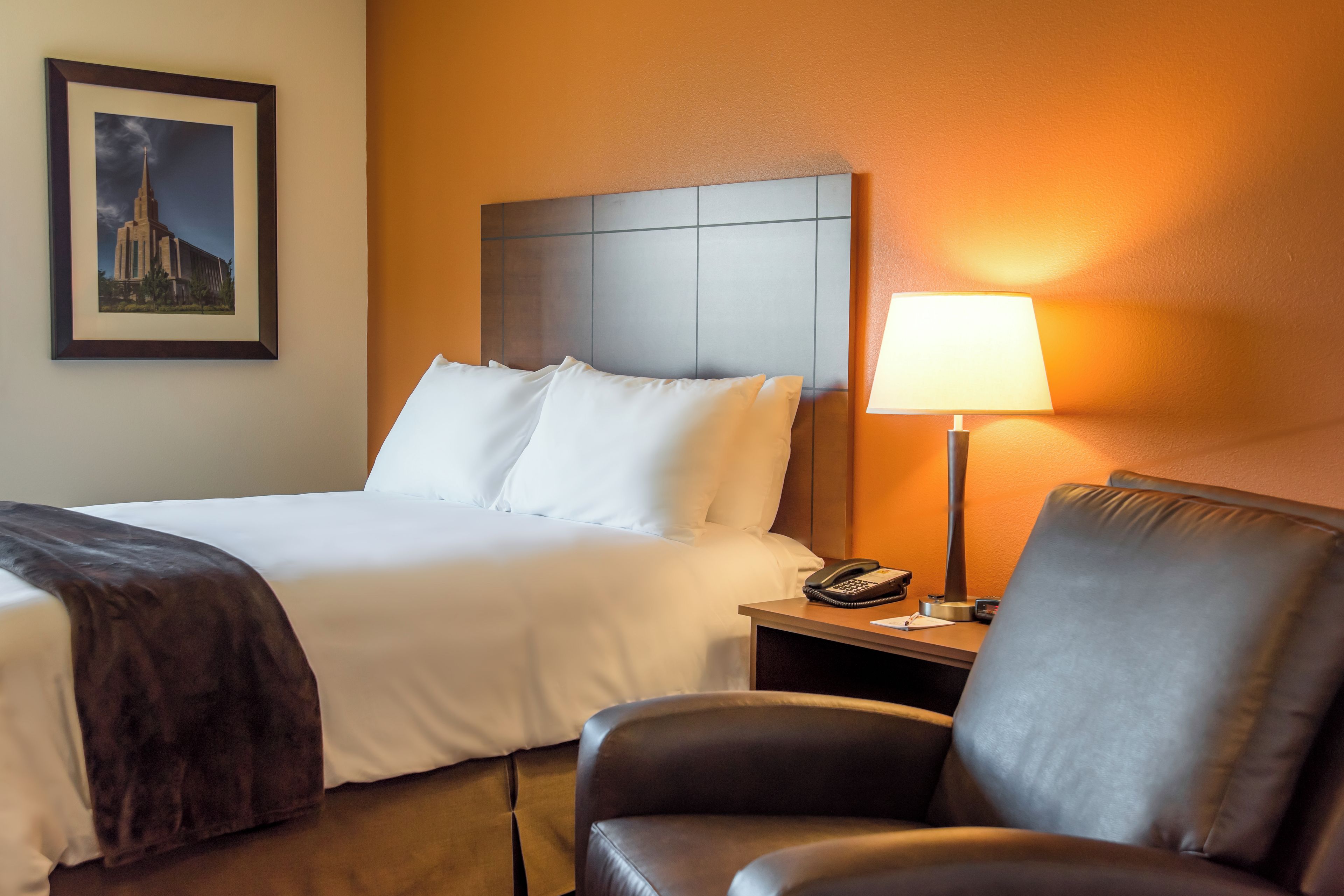 Une chambre d’hôtel avec un lit, une chaise, une table de chevet avec une lampe et un tableau encadré au mur.