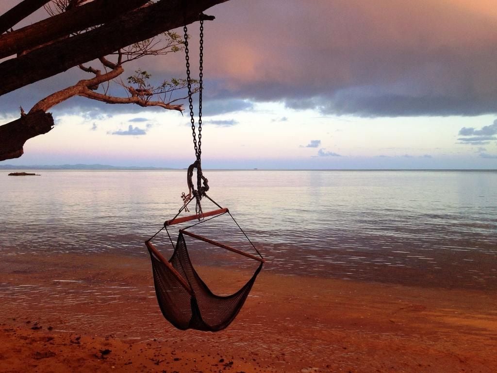 A hammock hanging from a wooden structure over a beach with calm water and a cloudy sky.
