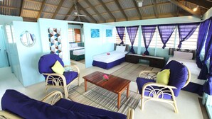 A room with a high ceiling, wooden beams, a bed with a blue bedspread, two chairs with blue cushions, a wooden table, and a window with purple curtains. - Funky Fish Beach Resort (Malolo Island)