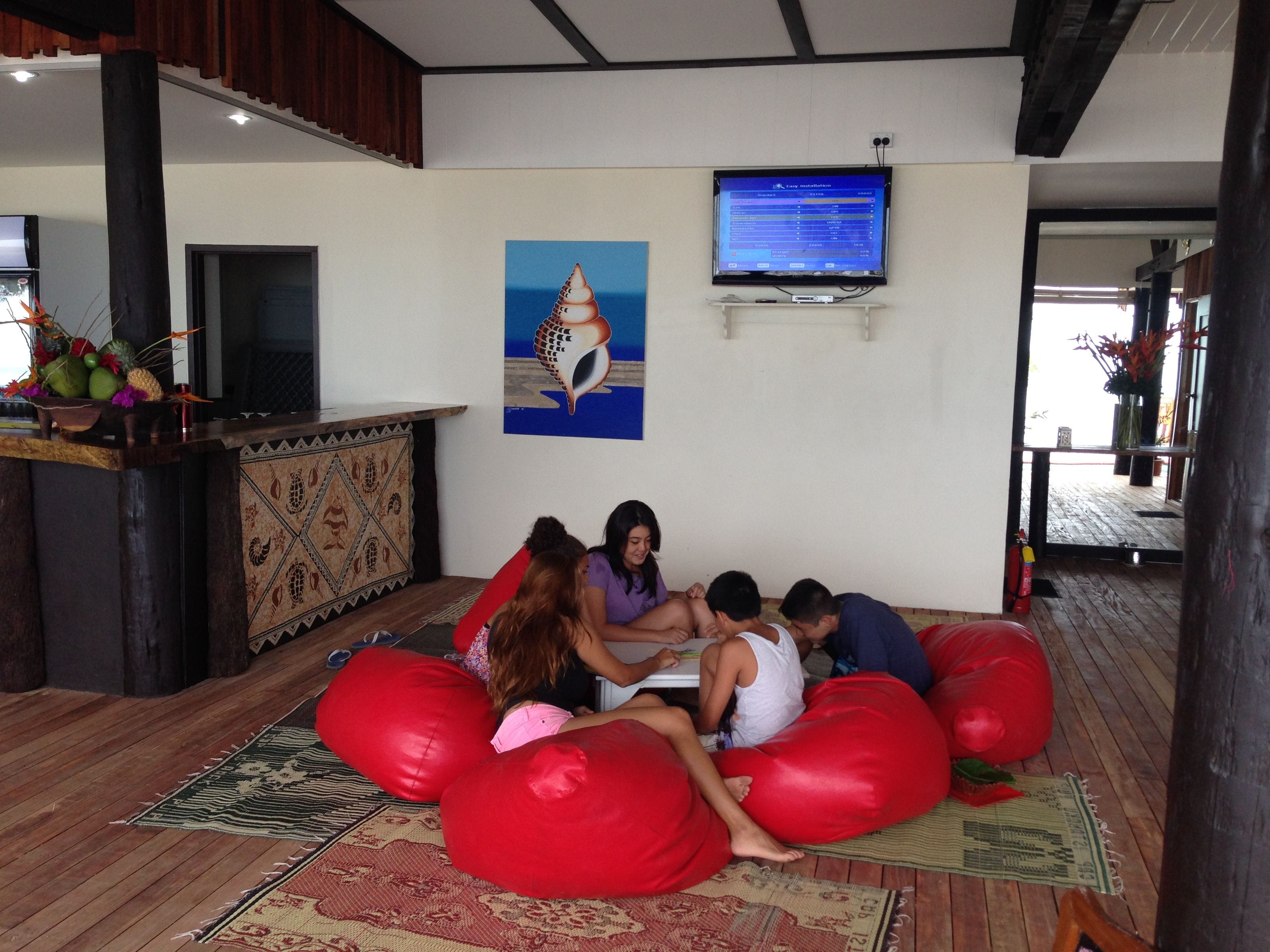 A group of people sitting on red bean bags in a room with a wooden floor, a television on the wall, and a painting of a shell.