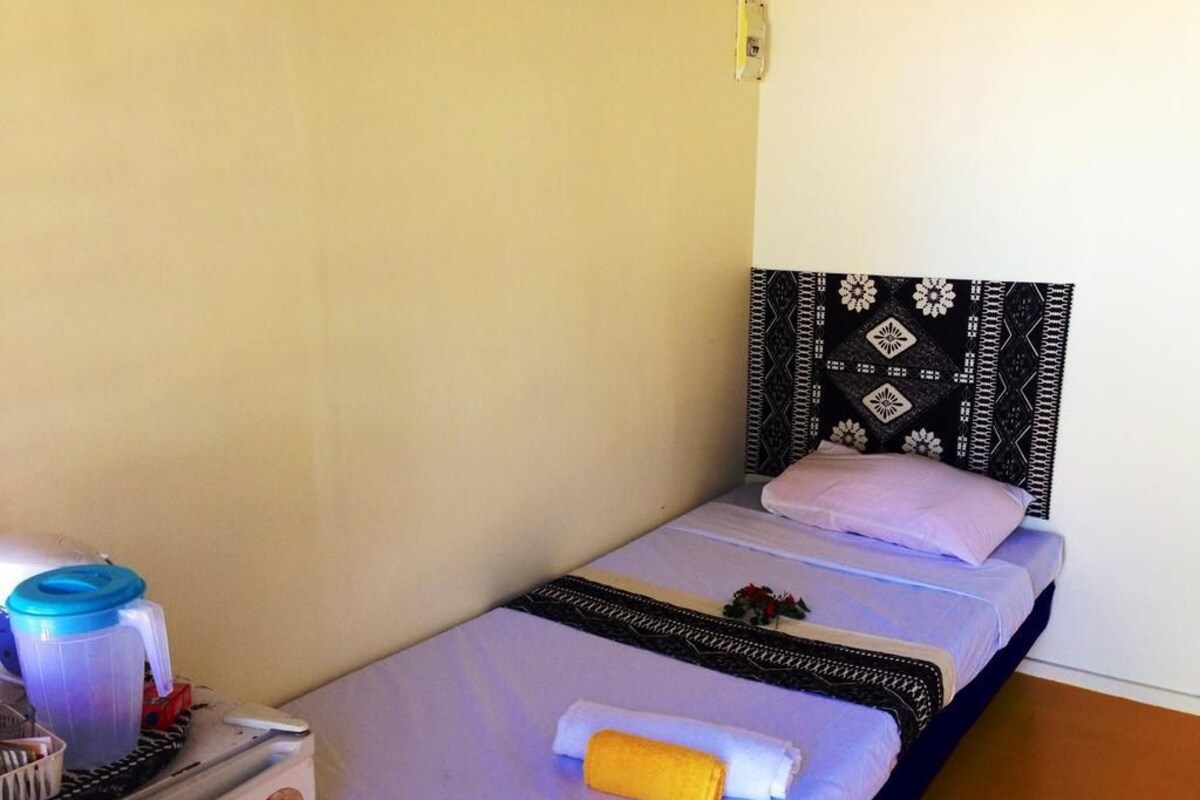A single bed with a patterned headboard, a small table with a blue container, and a white towel on the bed.