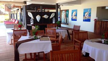 A covered outdoor dining area with wooden tables and chairs, a central decorative wall with shells, and a view of the sea.