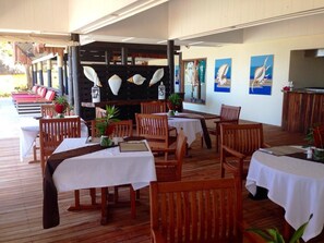 Een overdekt terras met houten tafels en stoelen, een decoratieve muur in het midden met schelpen, en uitzicht op de zee.
