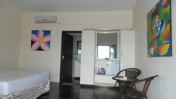 A room with a bed, a chair, a table, a refrigerator, and a door leading to a kitchen.