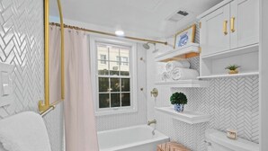 Une salle de bain avec un mur orné d’un motif en chevrons, une baignoire blanche, un tabouret en bois et une fenêtre donnant sur les bâtiments environnants.