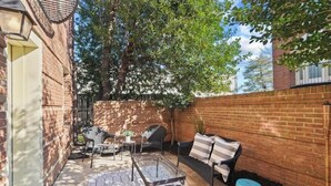 Une terrasse en briques avec des meubles d’extérieur, notamment une table et des chaises à plateau en verre, ainsi qu’une petite table agrémentée d’une plante.