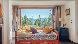 Ein Schlafzimmer mit großem Fenster, einem Bett mit bunter Bettwäsche, einem Nachttisch und einer Lampe.