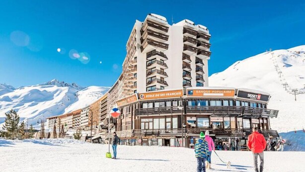 Un hôtel à plusieurs étages, des panneaux d’indication menant à la station de ski, et une montagne enneigée en toile de fond.