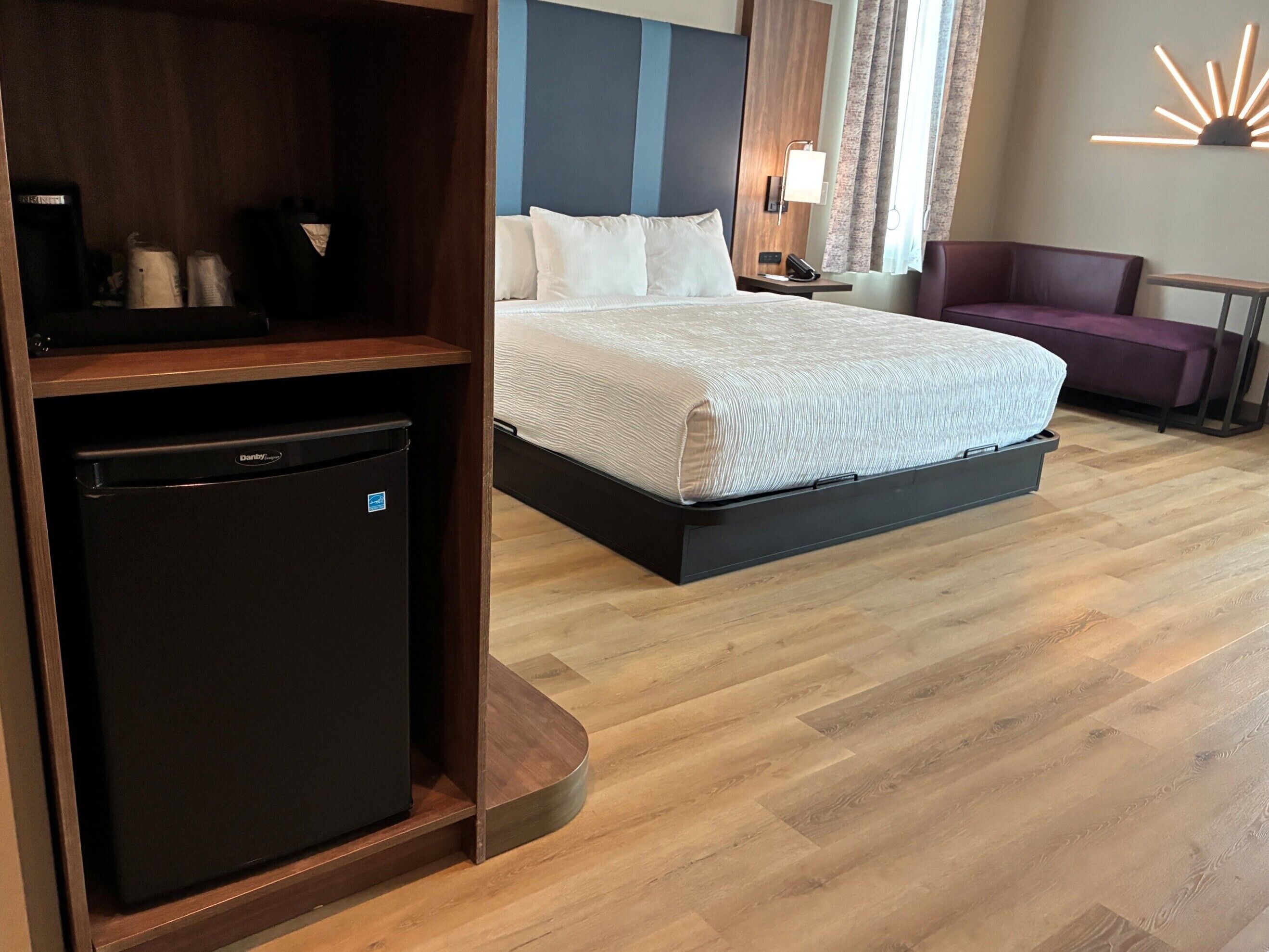 Habitación de hotel con cama, escritorio con computadora, refrigerador pequeño, silla y lámpara.