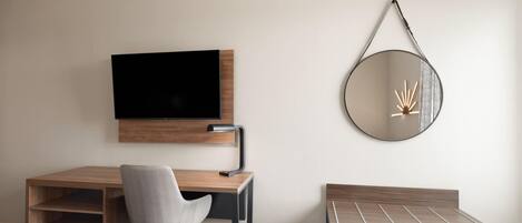 Een moderne hotelkamer met een houten bureau, een stoel, een televisie, een ronde spiegel en een lichtblauwe koffer.
