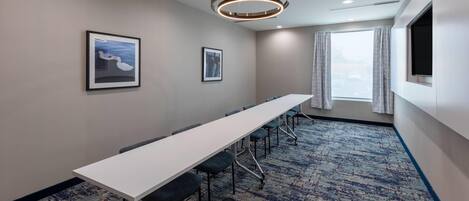 A conference room with a long table, chairs, framed pictures on the wall, a large window with curtains, and a patterned carpet.